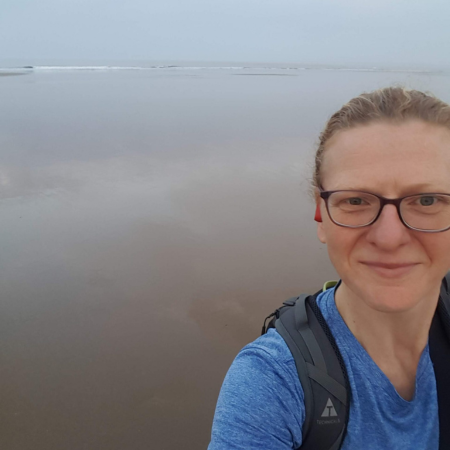 Celia is standing on the beach. She is wearing a blue t-shirt and a rucksack. Celia is wearing glasses.